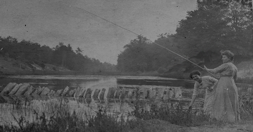 Black and white photo of a lady fishing by a river.