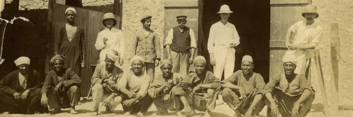 Row of Egyptian men sitting on ground with row of men in white suits standing behind.