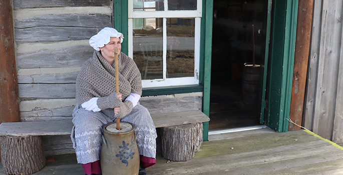 Lady dressed in early settler clothing with a bonnet on churning butter.