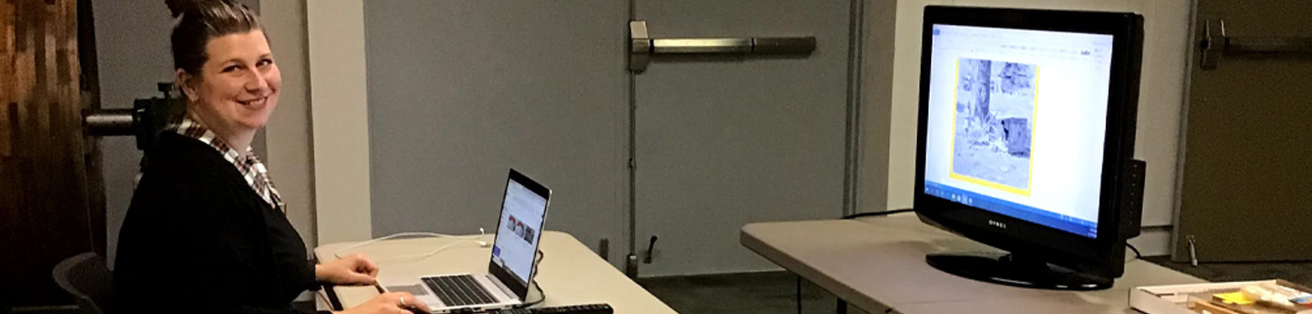 Lady sitting at desk with a computer and tv.